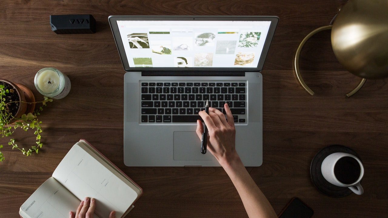 Person typing one-handed on a laptop while holding a notebook open with their other hand